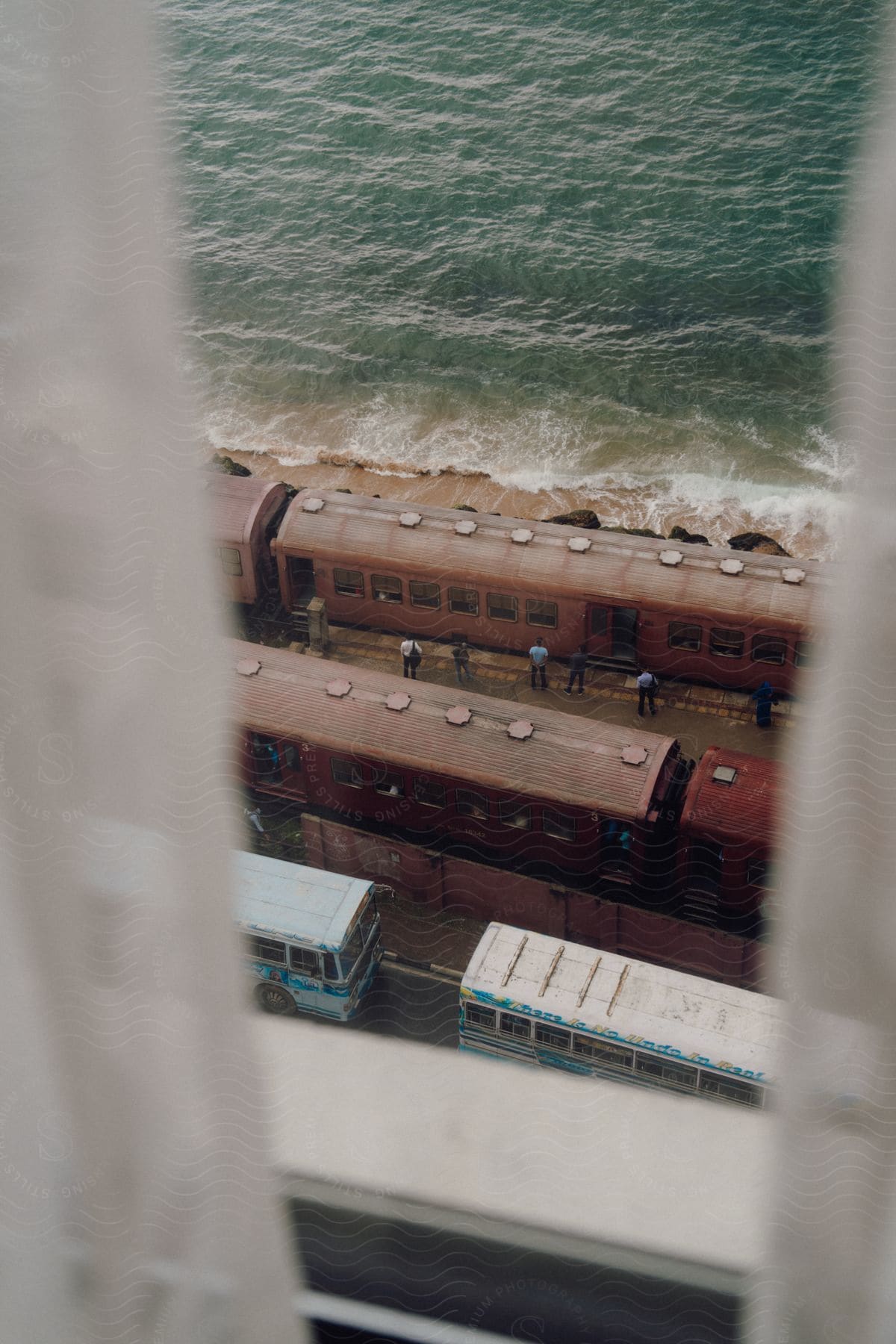 An elevated view through a narrow gap between the curtains of a window reveals a red train with people on it, parked next to the sea.