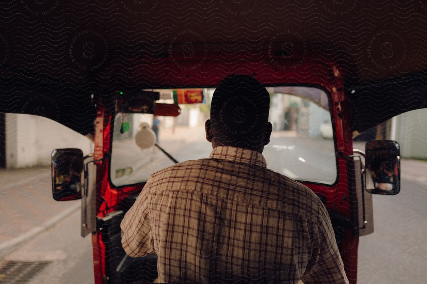 View from the back of a red tuk-tuk with the driver in a plaid shirt navigating through a quiet street.