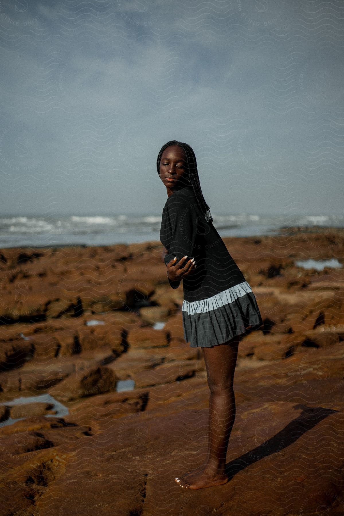 A woman with dreadlocks, wearing a black jacket and a gray skirt, stands barefoot on a rock on the beach near the wavy sea.