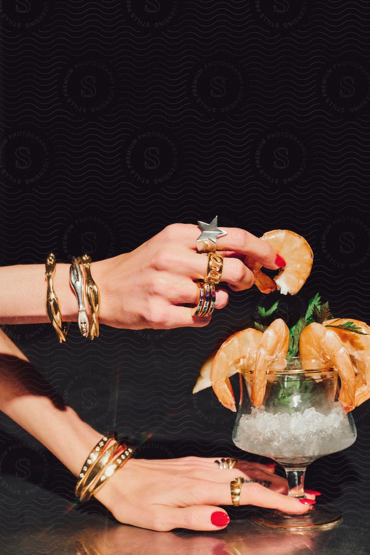 Two hands with various jewels holding a shrimp in a glass filled with ice.