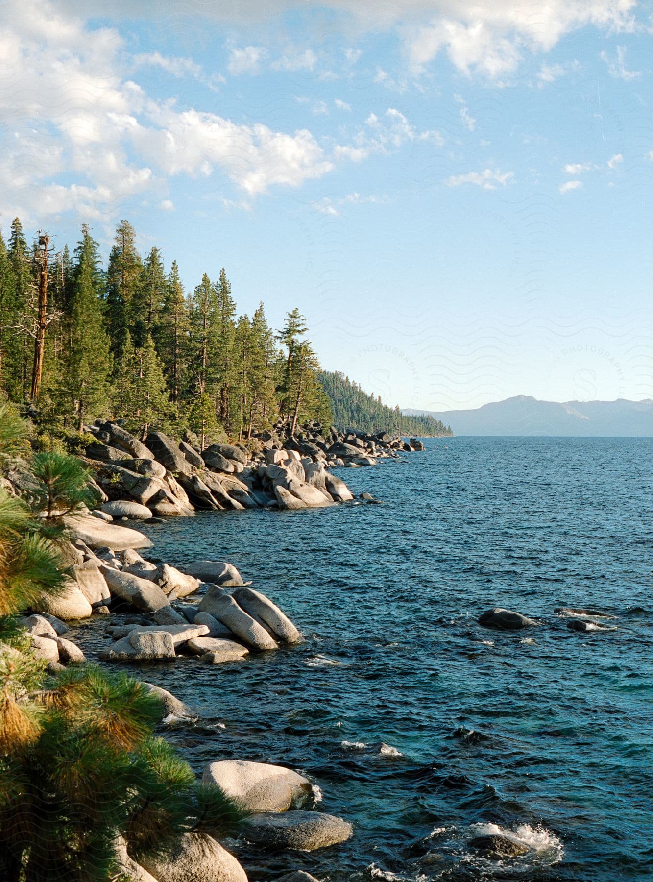 A serene scene of a lake with blue water, surrounded by a rocky shore and pine trees, with mountains in the background.