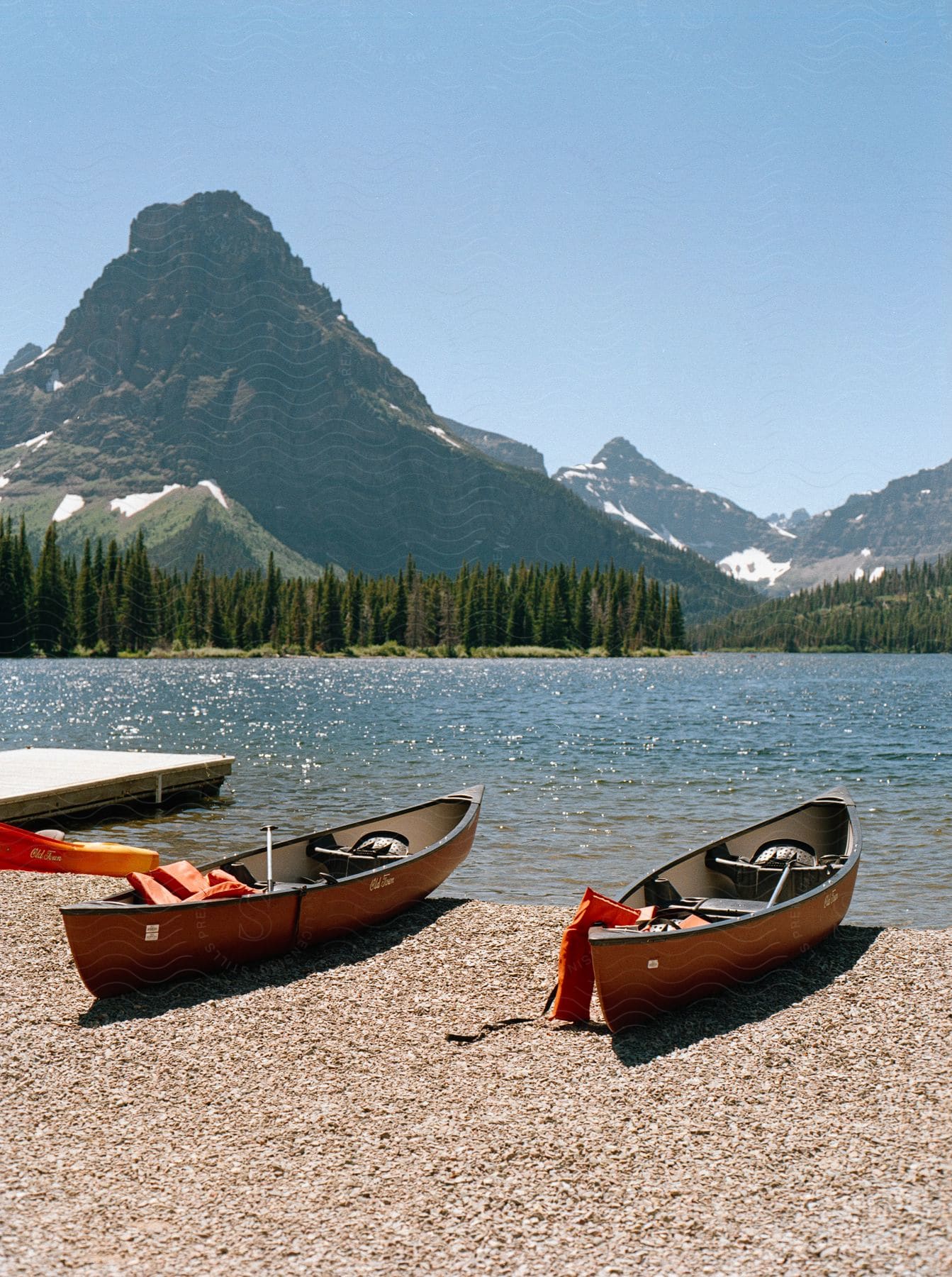 Two canoes rest on a pebble shore beside a serene lake with a mountain and clear sky in the background