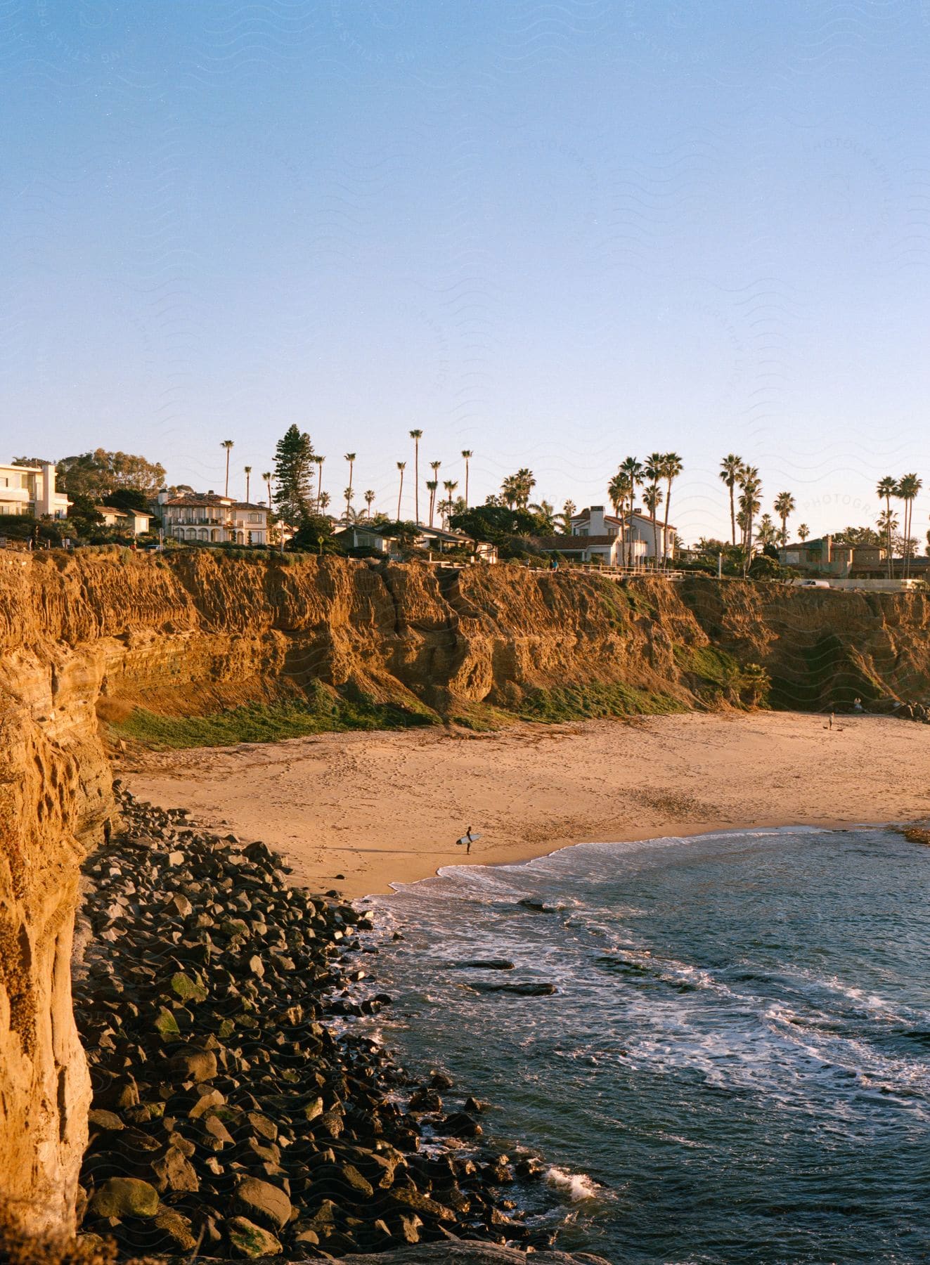 A person walks on a beach with palm trees and houses on a cliff above and ocean waves at the shore