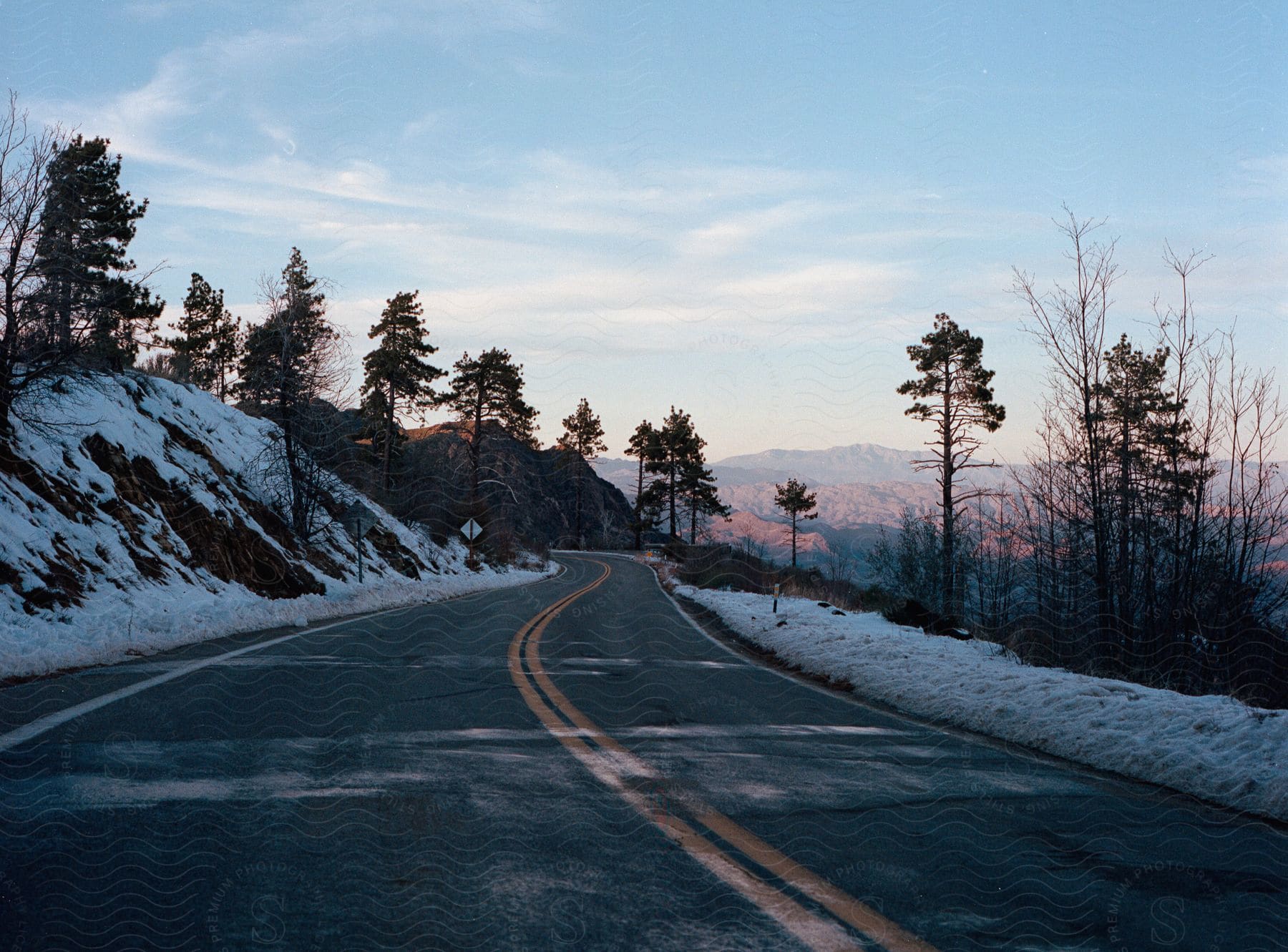 A winding road surrounded by snow and trees, with mountain scenery in the background at dusk.