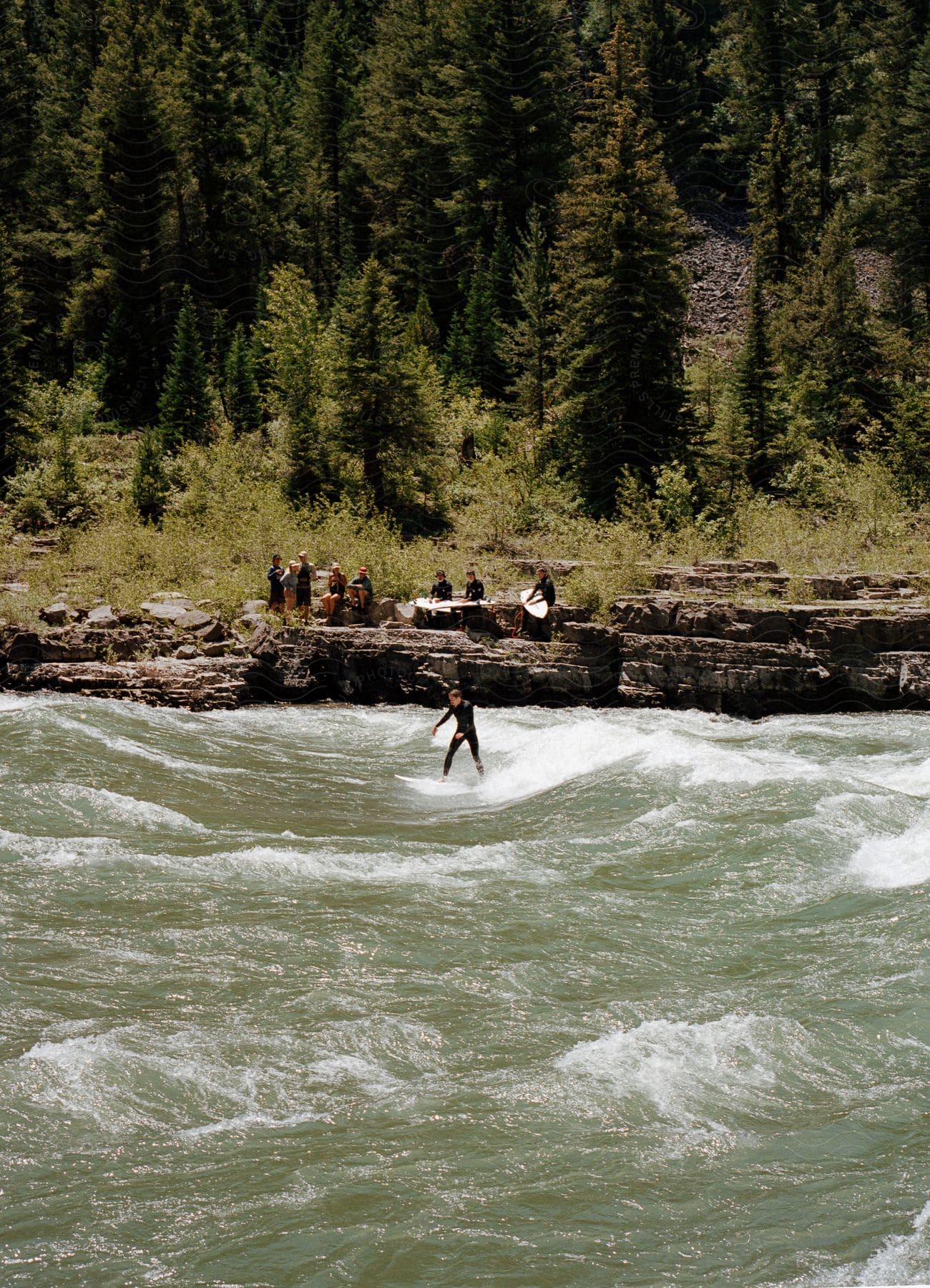 A person is surfing in the river while others are watching from the bank during the daytime.