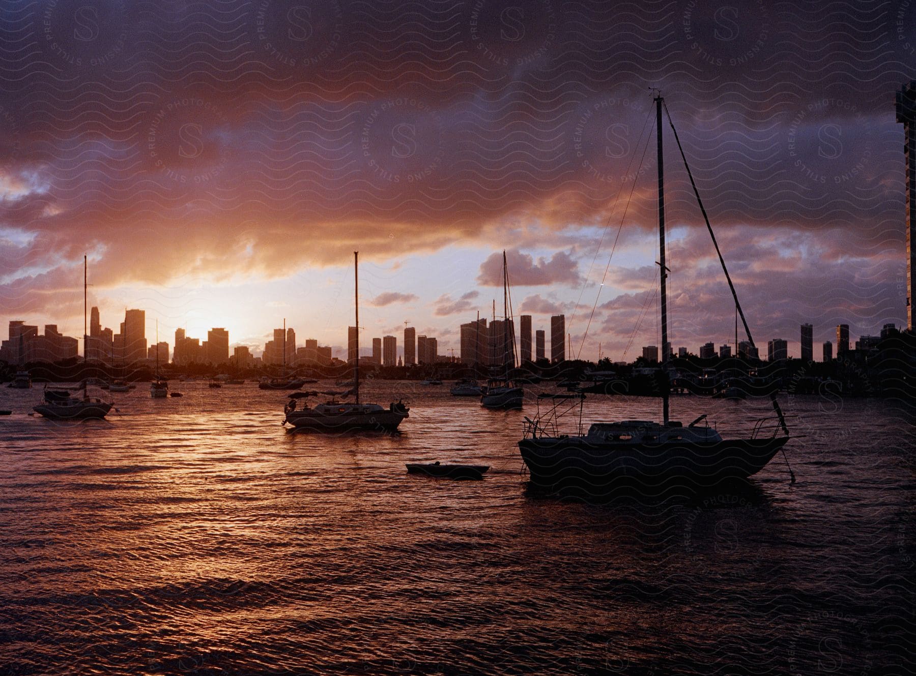 Boats sail in the water as the sun shines on the horizon behind city buildings and skyscrapers