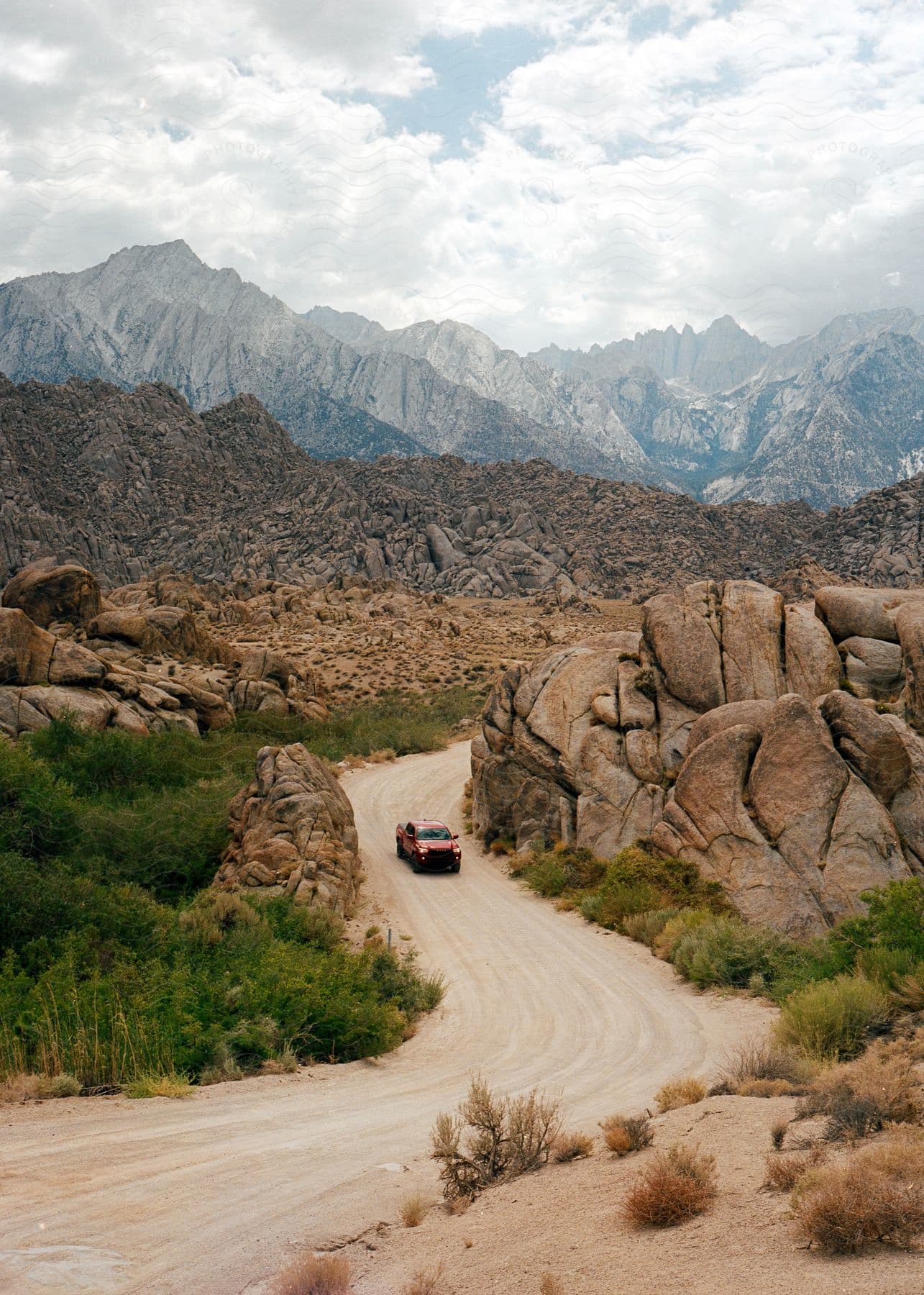 Vehicle driving on a winding dirt road through a rocky desert landscape with mountains in the background.