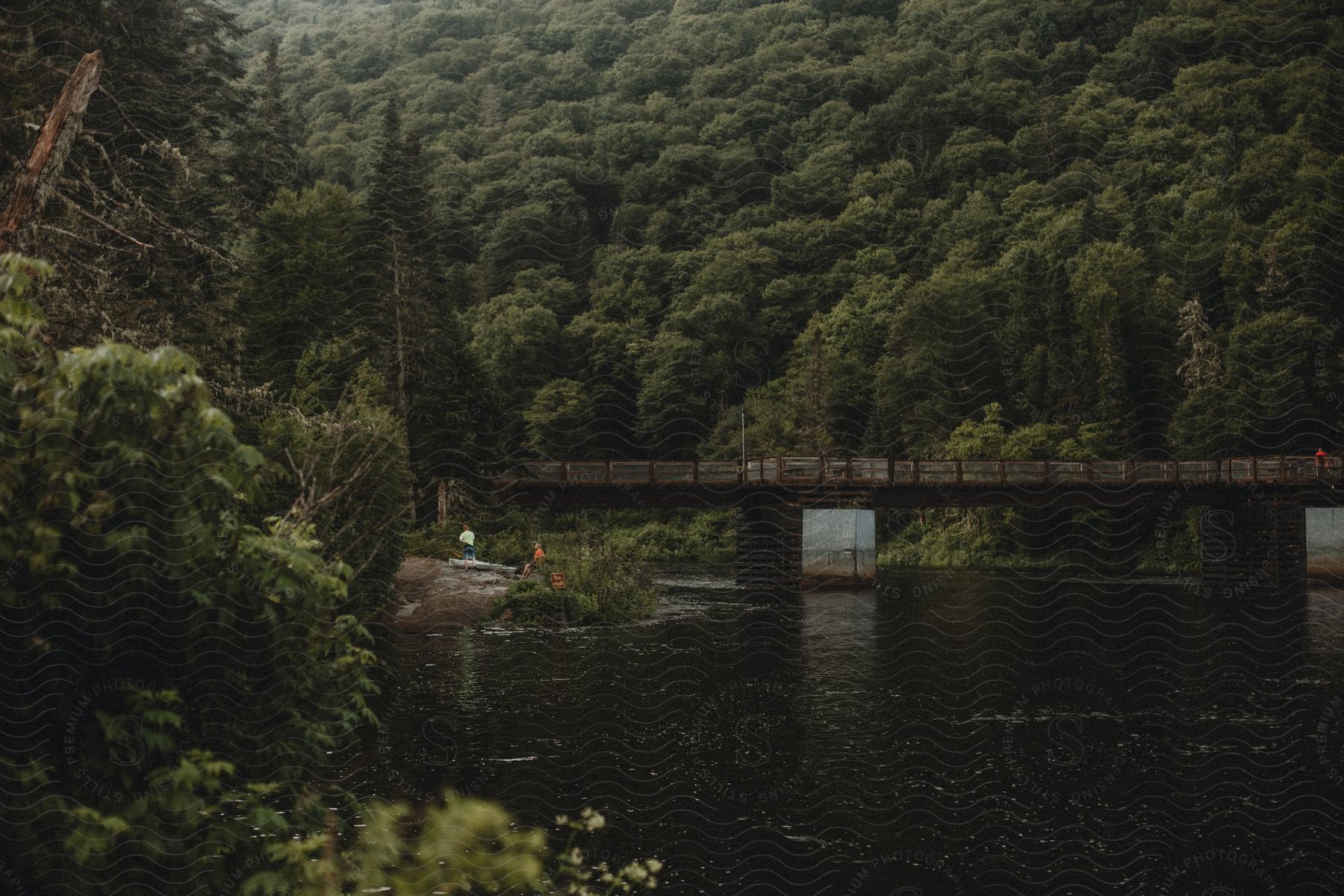 Two men stand on a riverbank near a wooden bridge with a railing spanning across a forested area on a mountain slope.