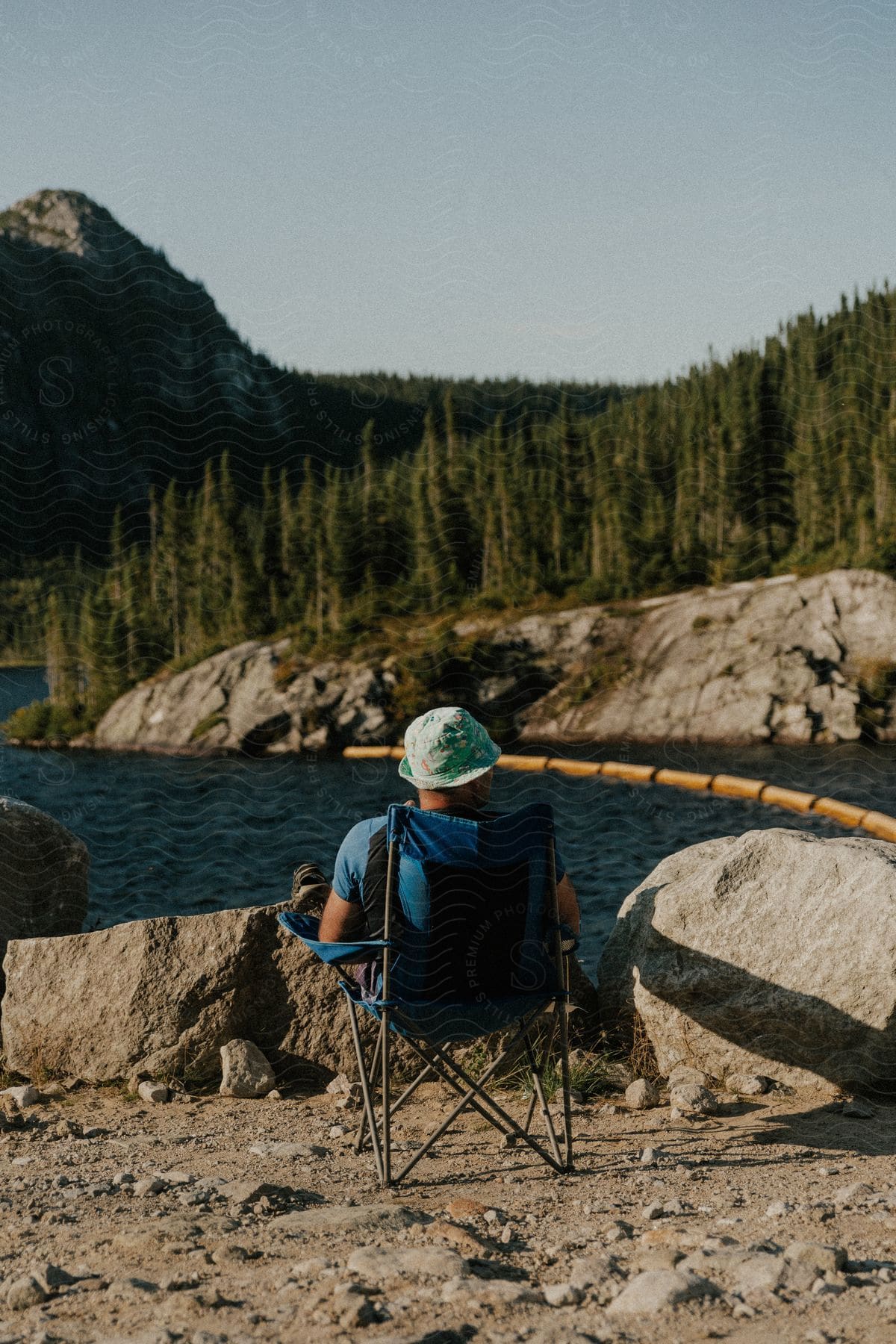 A man sits alone beside a rocky river bank during the daytime, surrounded by trees.