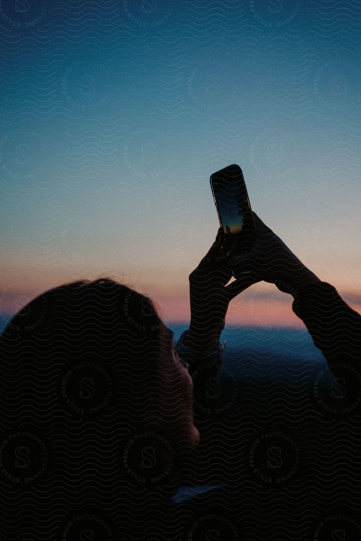 A woman holding a smartphone up takes photos of the sunset sky in the field.