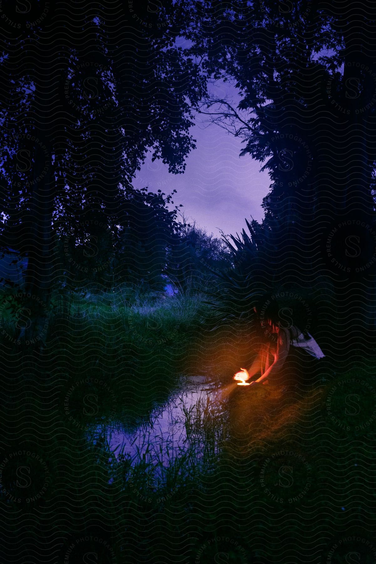 Woman sits at side of stream in woods with a light.