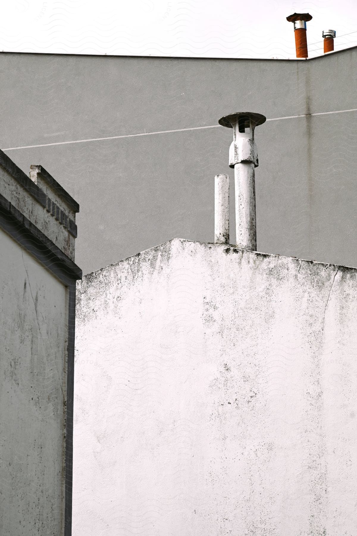 A white weathered wall with a pipe and cap next to a grey building with two chimneys