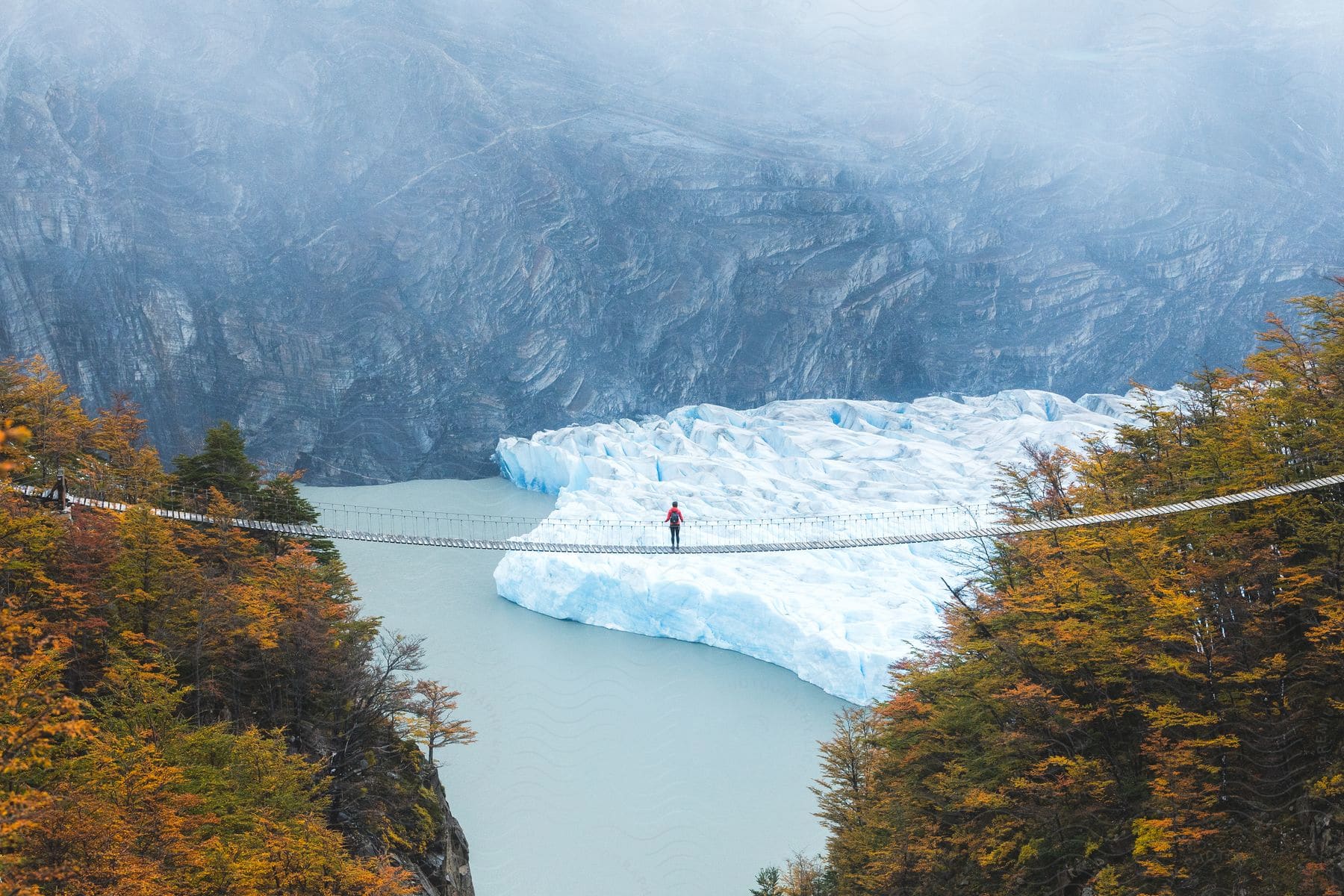 A lone figure traverses a suspension bridge over a river, framed by mountains, a glacier, and autumn foliage