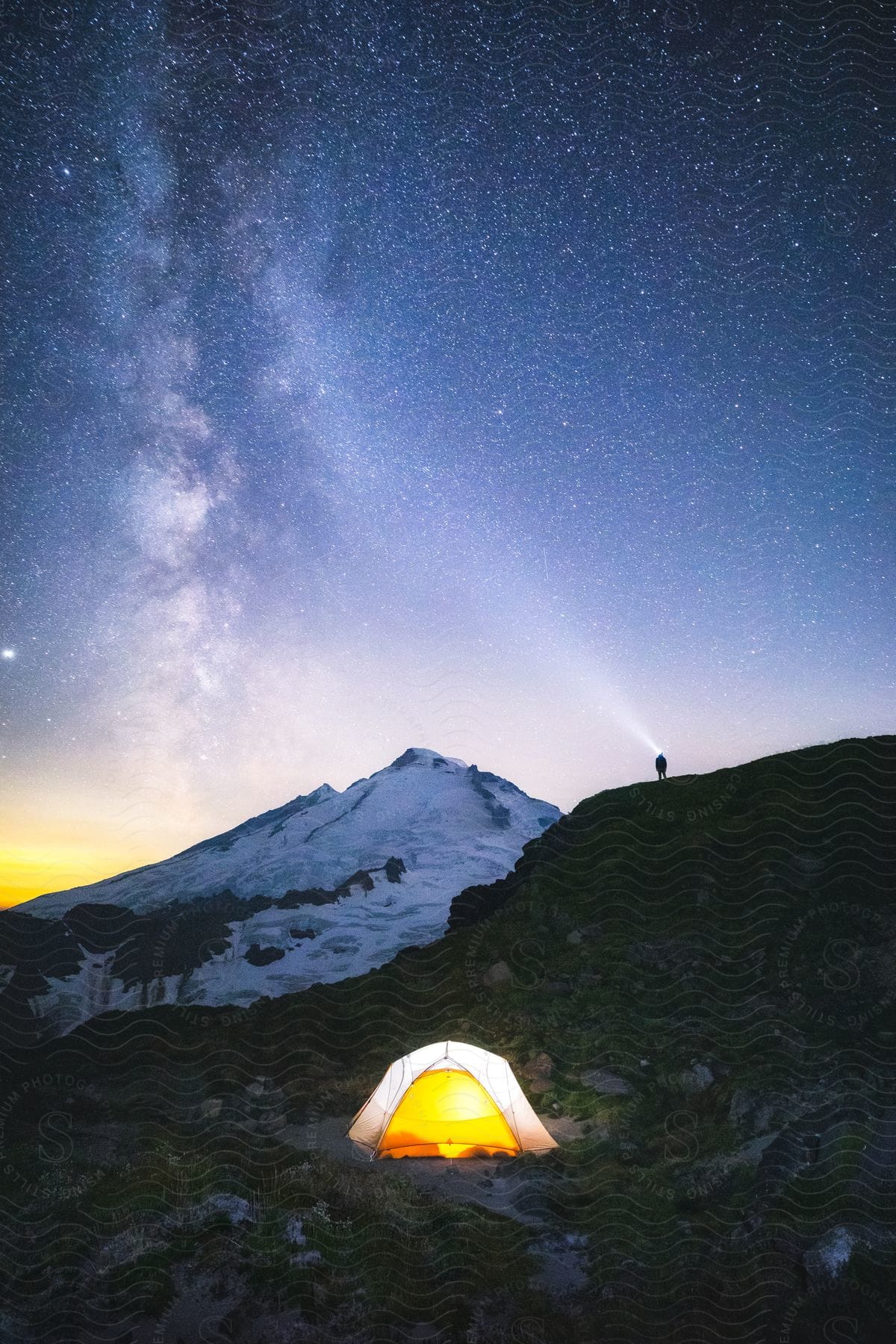A person sets up a tent on top of a mountain at night, looking into the sky with a flashlight.