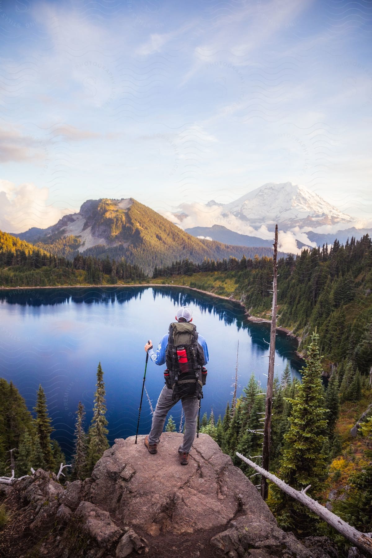 A hiker, with a backpack and trekking poles, stands on a rock, gazing out at the serene sea and the snow-capped mountain landscape.