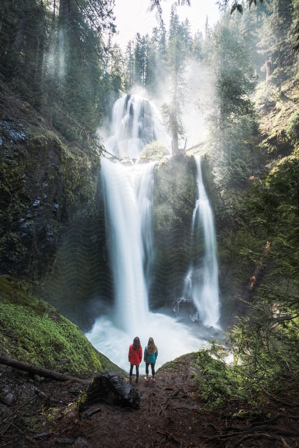 Waterfall with multiple waterfalls surrounded by dense vegetation and two women on a cliff enjoying the view.