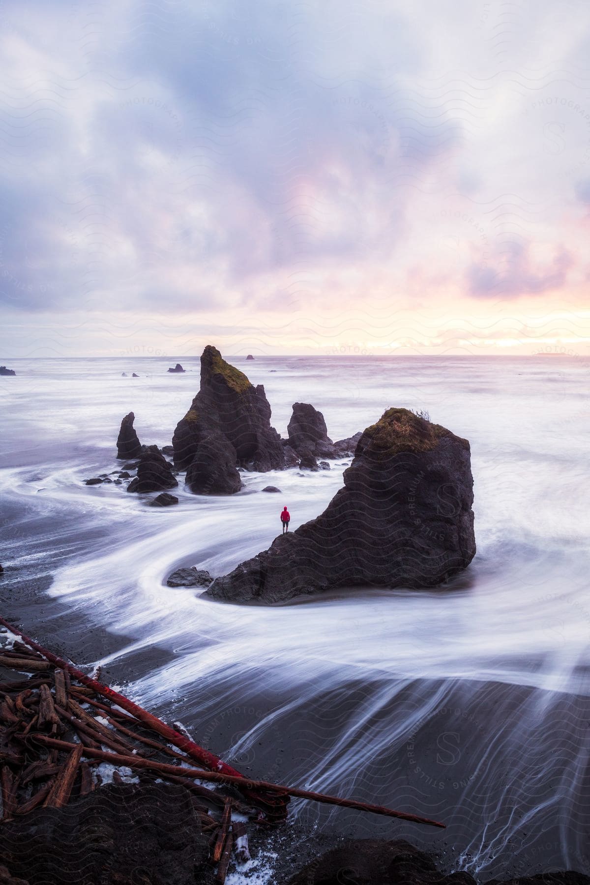 A man wearing a red jacket stands on a rocky outcropping, gazing out at the vast expanse of the Pacific Ocean.