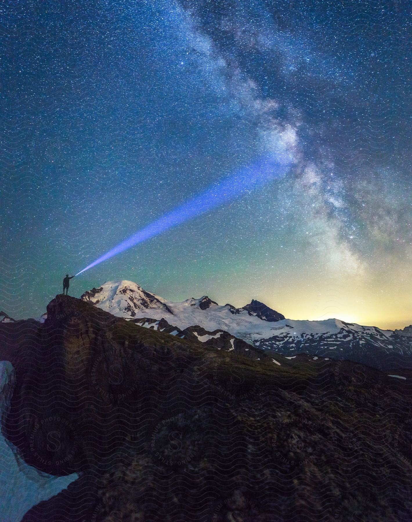 Person shines a blue flashlight towards the Milky Way while standing on a rocky peak with snow-covered mountains in the background.