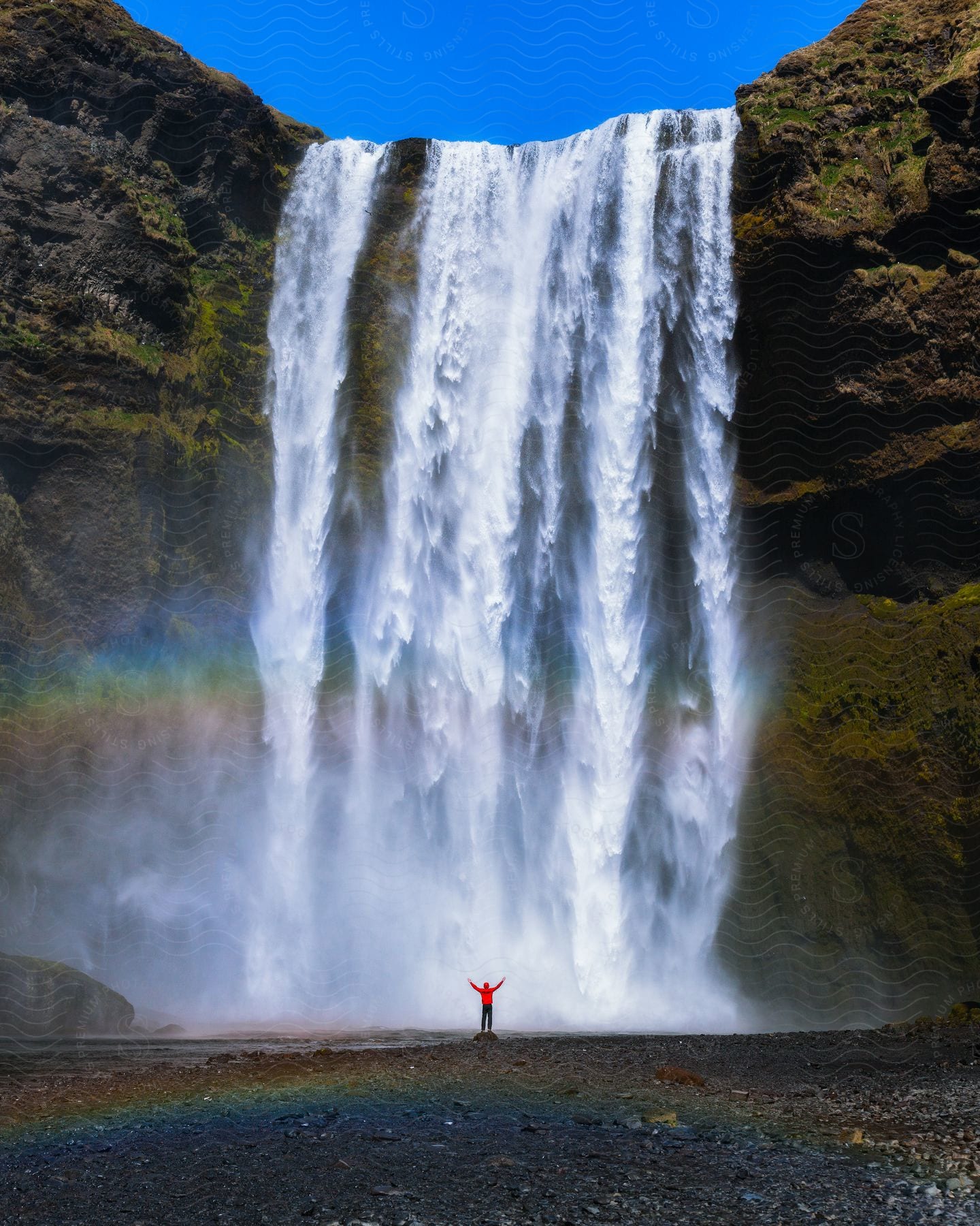 A person wearing a red jacket stands at the base of a giant waterfall.