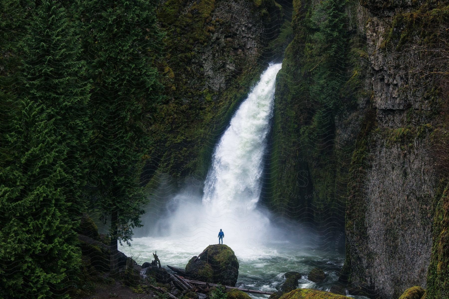 A person stands on top of a large boulder in front of a rushing waterfall.