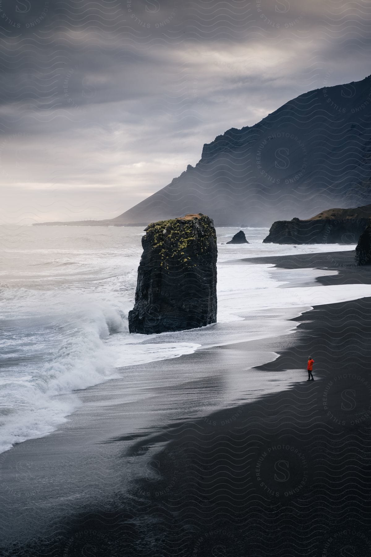 Person in an orange jacket standing on a black sand beach next to a tall sea stack with waves crashing against it, and mist-covered cliffs in the background.