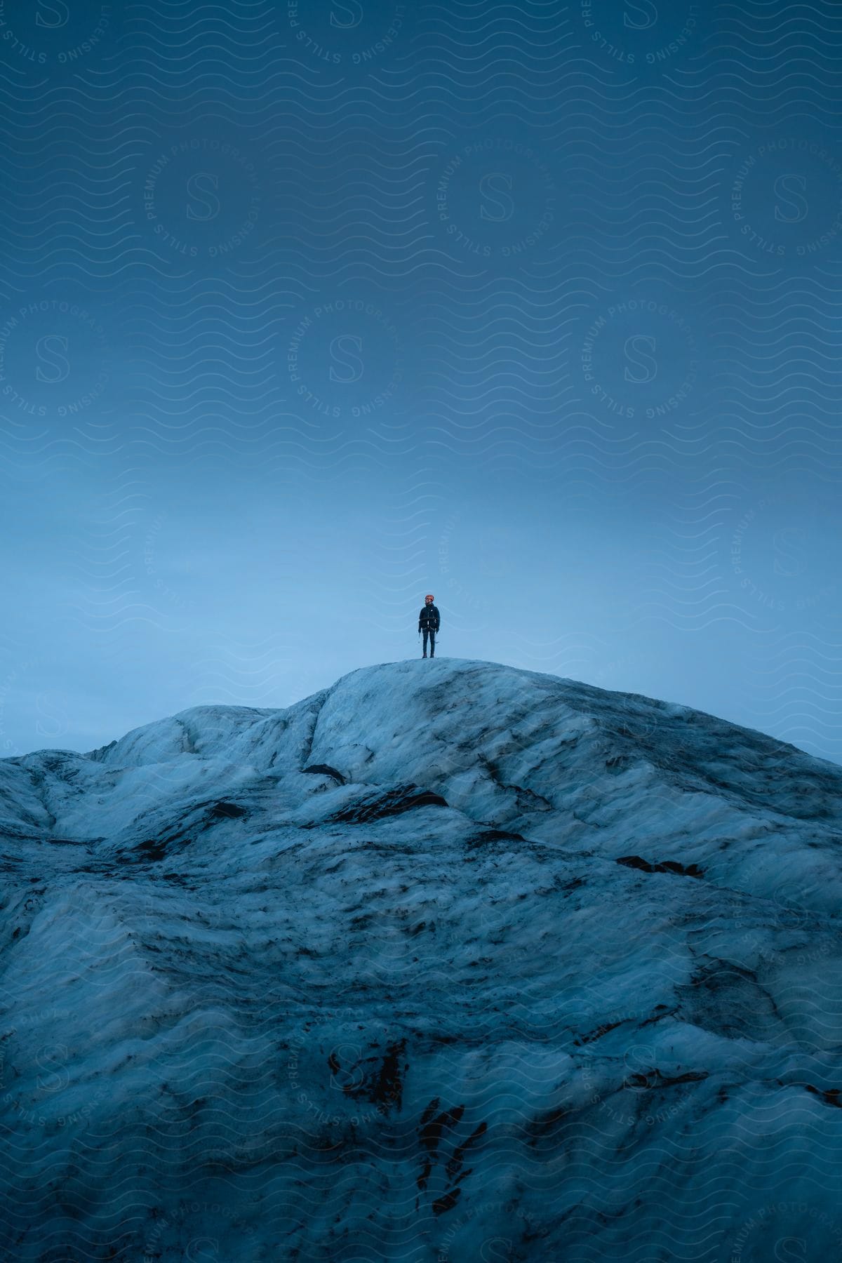 Stock photo of a natural landscape dominated by cold, blue tones, with a person standing at the top of a snow-covered ridge under a dark blue sky.