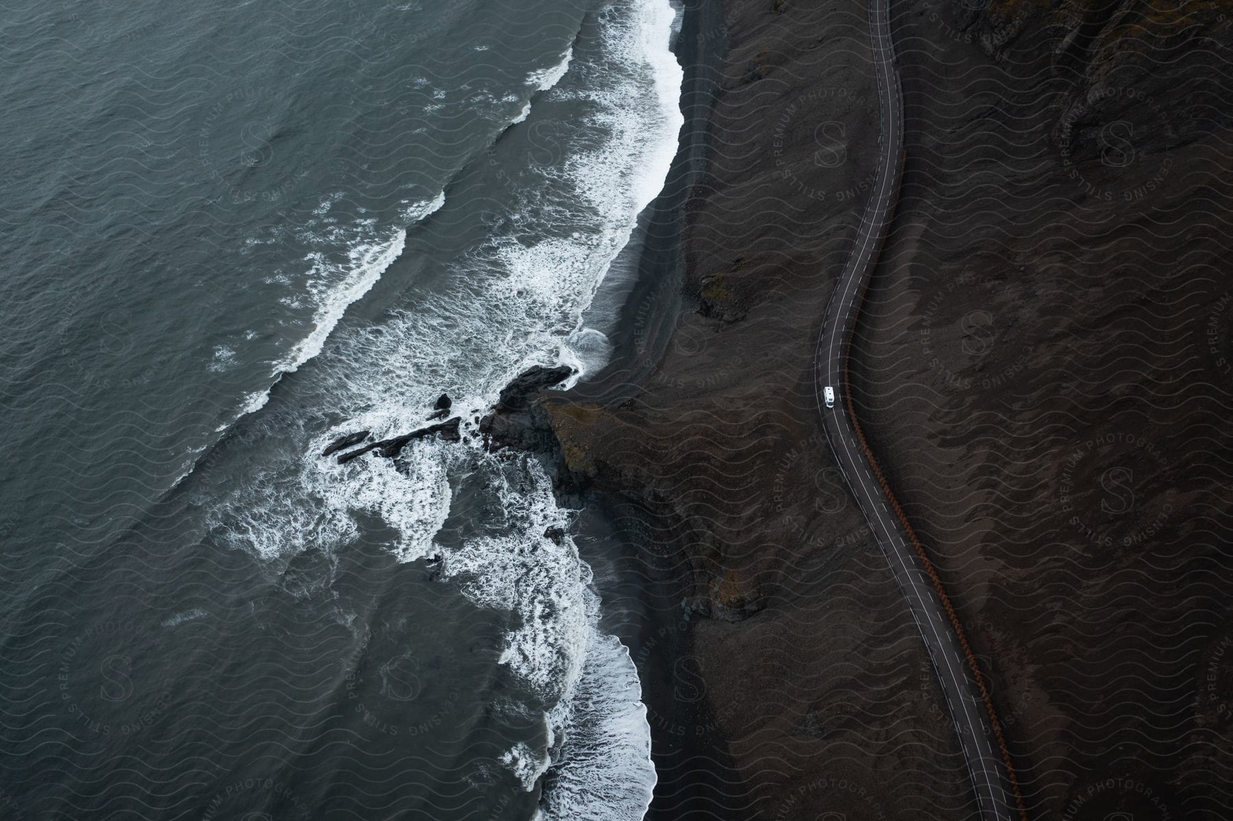 An aerial view captures a white van driving on a winding road near the ocean.