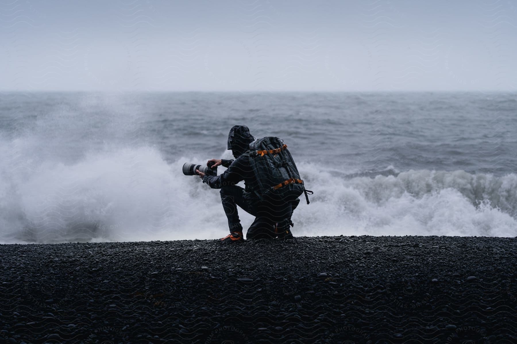 A photographer with backpack aiming camera at rough sea from rocky shore under gloomy weather
