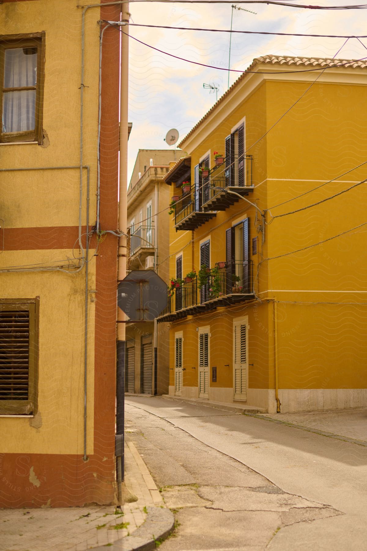 A bright yellow building with a black wrought iron balcony flanks a narrow street lined with cobblestones.