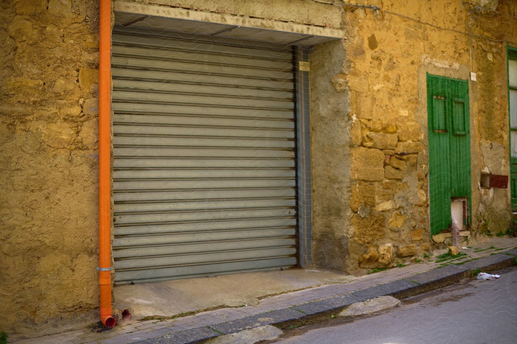 A closed grey shutter door beside a green wooden door on a worn wall with an orange drainpipe