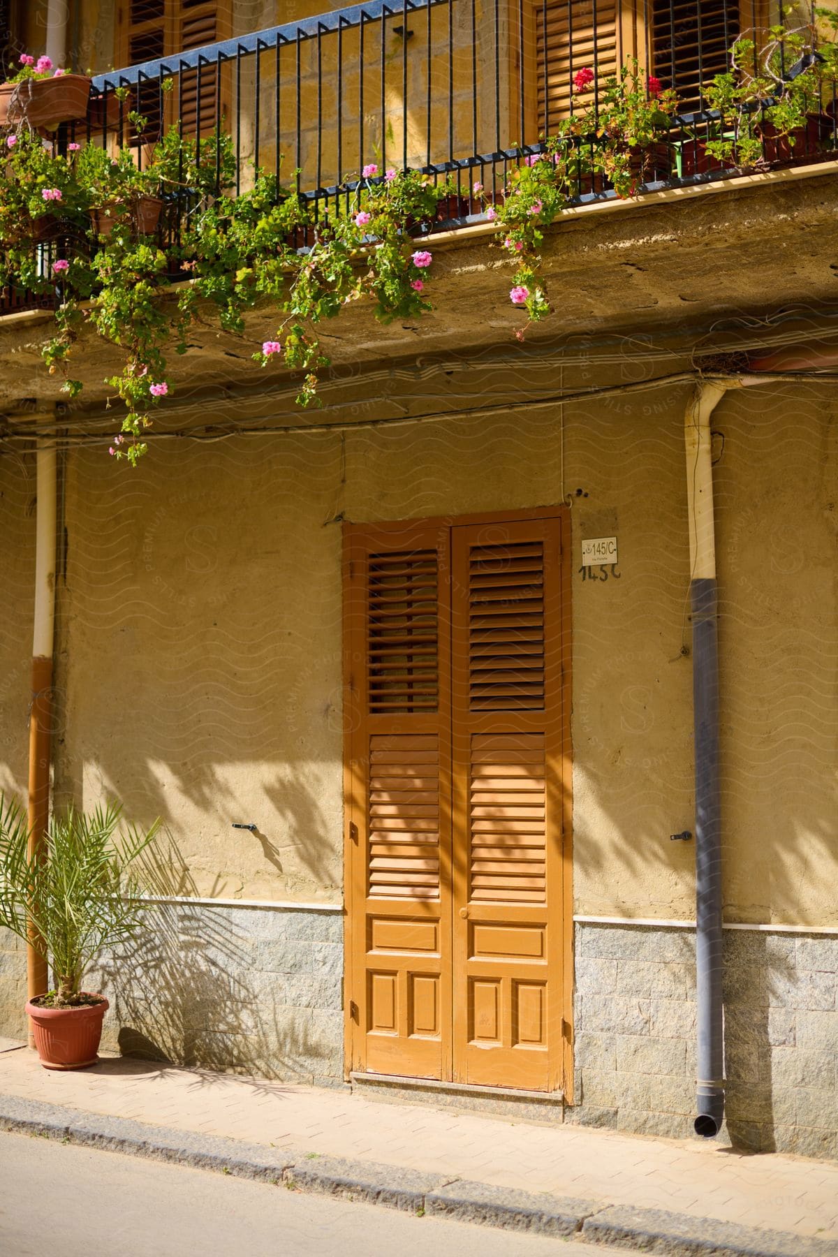 A building with a brown wooden door and a balcony overflowing with colorful flowers and plants stands elegantly beside a charming cobblestone street.