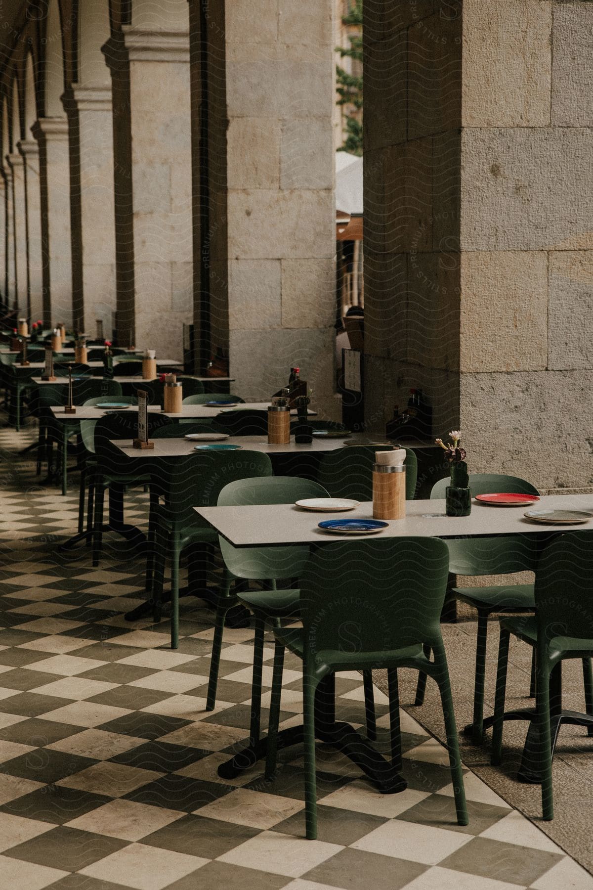 Outdoor café with green chairs and white tables on checkerboard flooring near stone columns