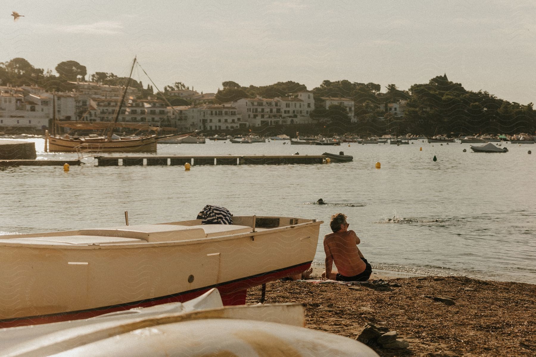 Man sitting on a sandy beach beside a rowboat, looking out over a calm harbor with two swimmers in front, and a town and hills in the background.