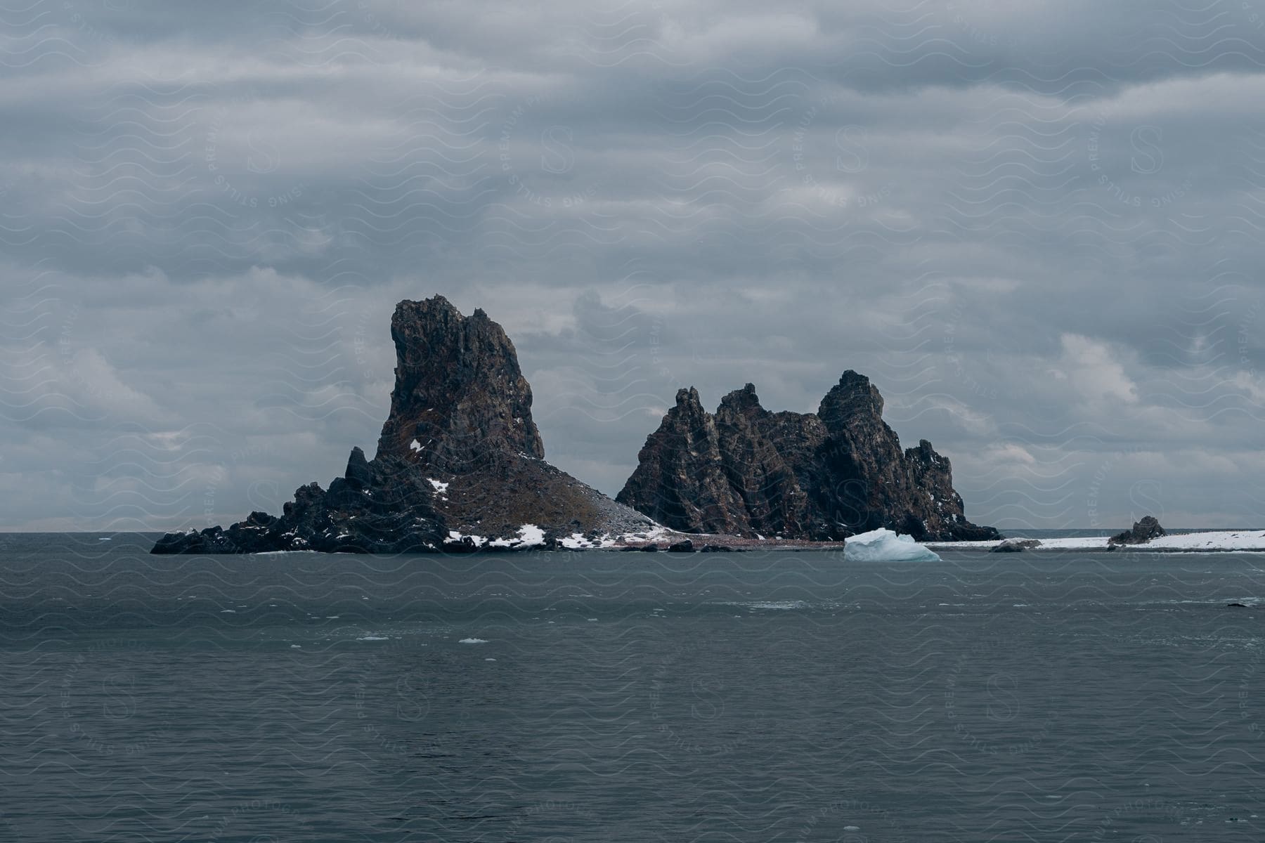 A rocky island with patches of snow is surrounded by a calm sea under a cloudy sky.