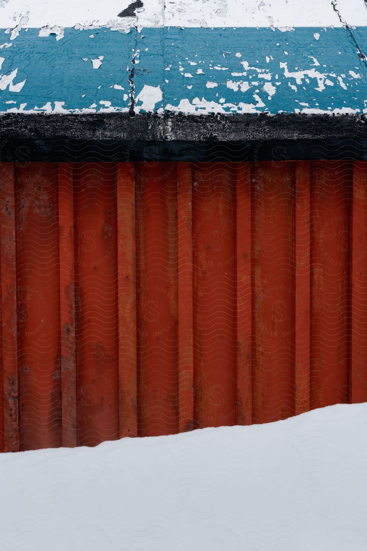 A weathered blue roof wall above a rusty red wall partially covered in snow