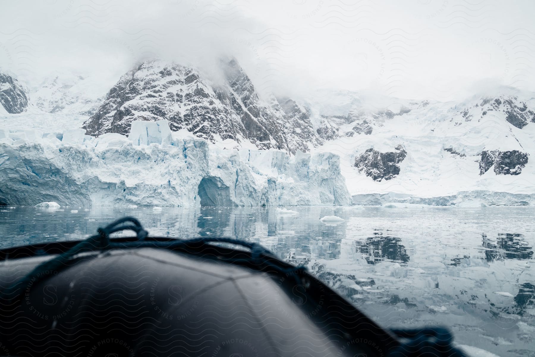 An impressive view of an icy landscape, seen from the perspective of someone in a boat. The black bow of the boat points towards the icy landscape, with calm waters reflecting the magnificent ice formations and snow-capped mountains.