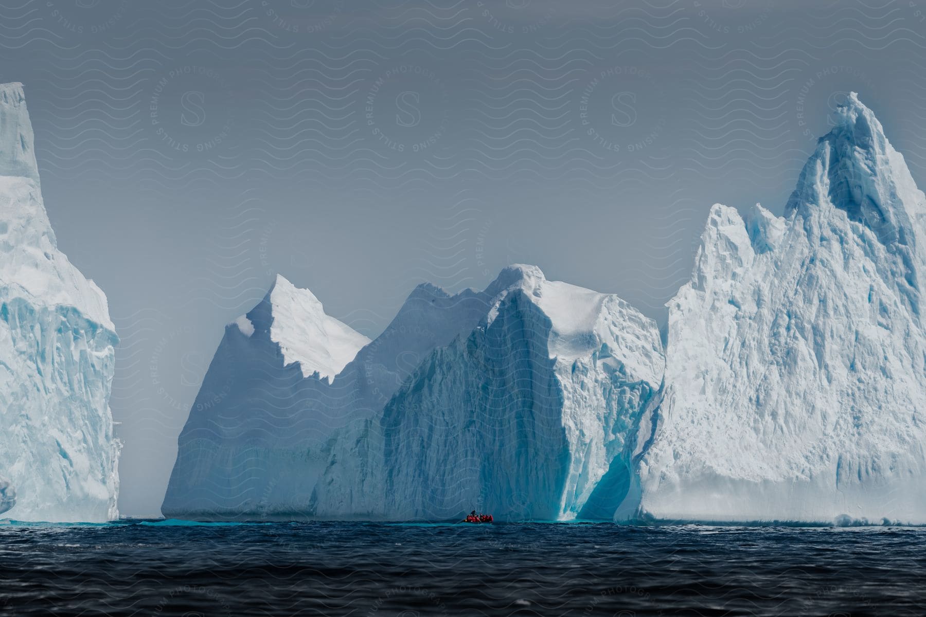 A small boat sails close to huge icebergs with jagged peaks in a cold and serene ocean setting.