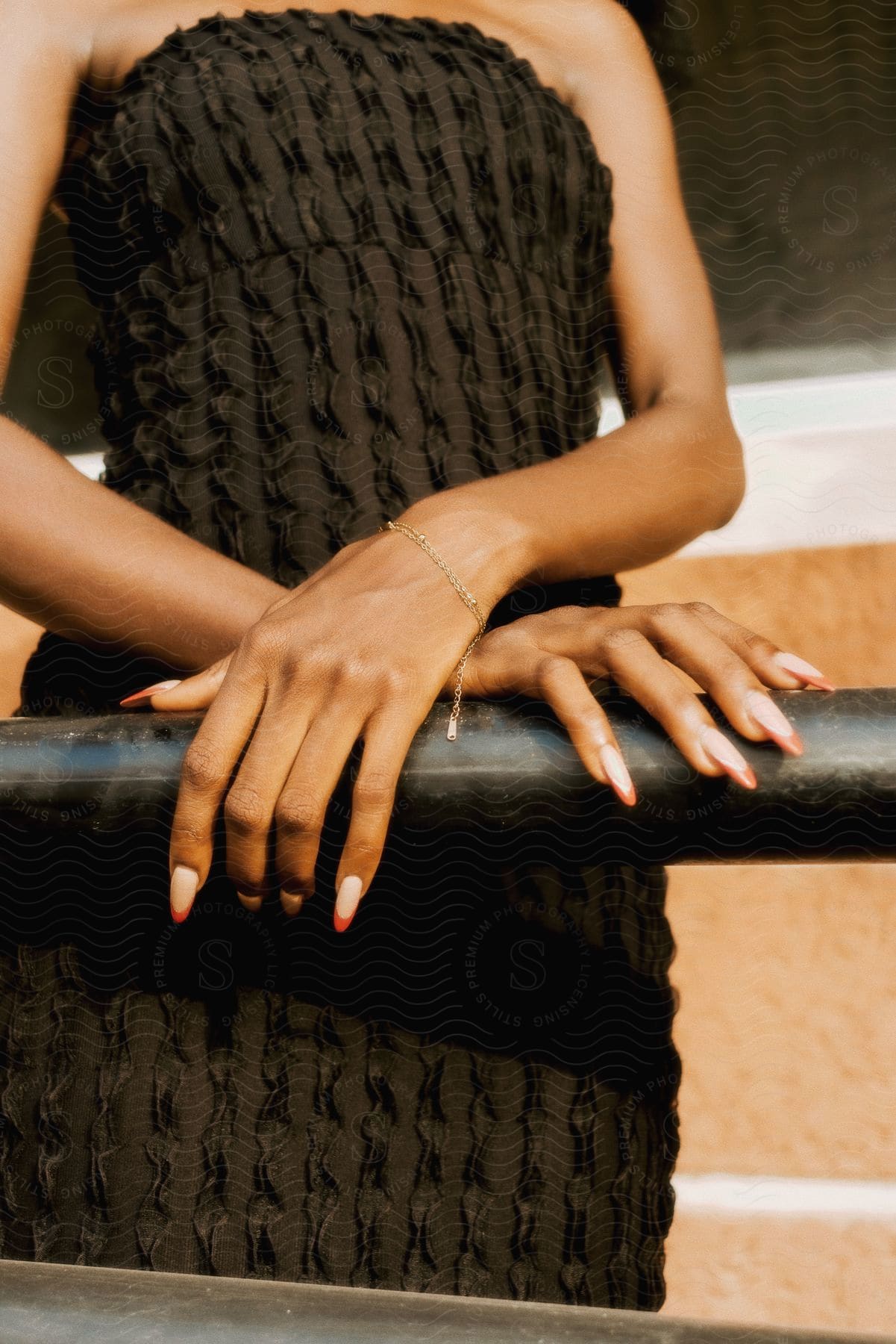 A woman in a textured black dress rests their hands with long nails on a rail