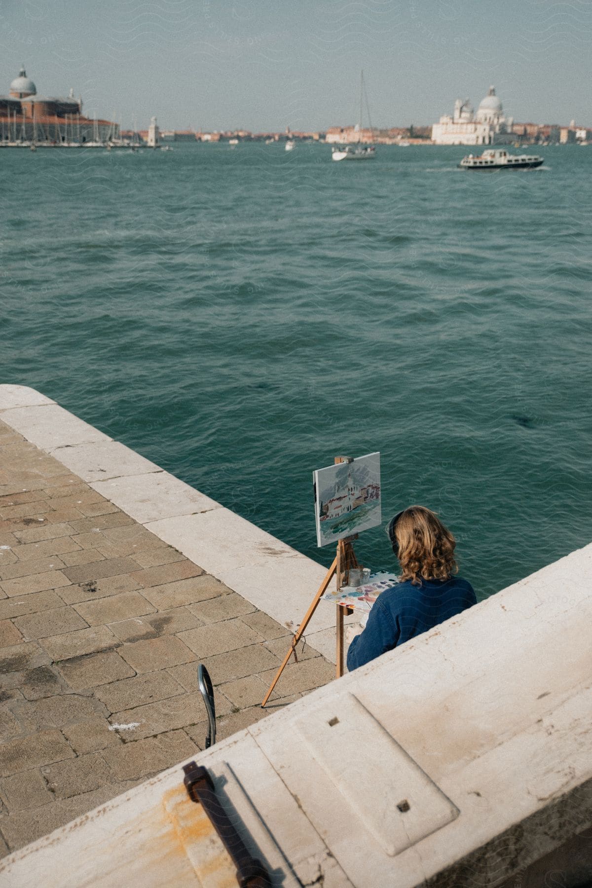 Stock photo of artist painting on an easel next to the water with a view of boats sailing and historic buildings in the distance under a clear sky.