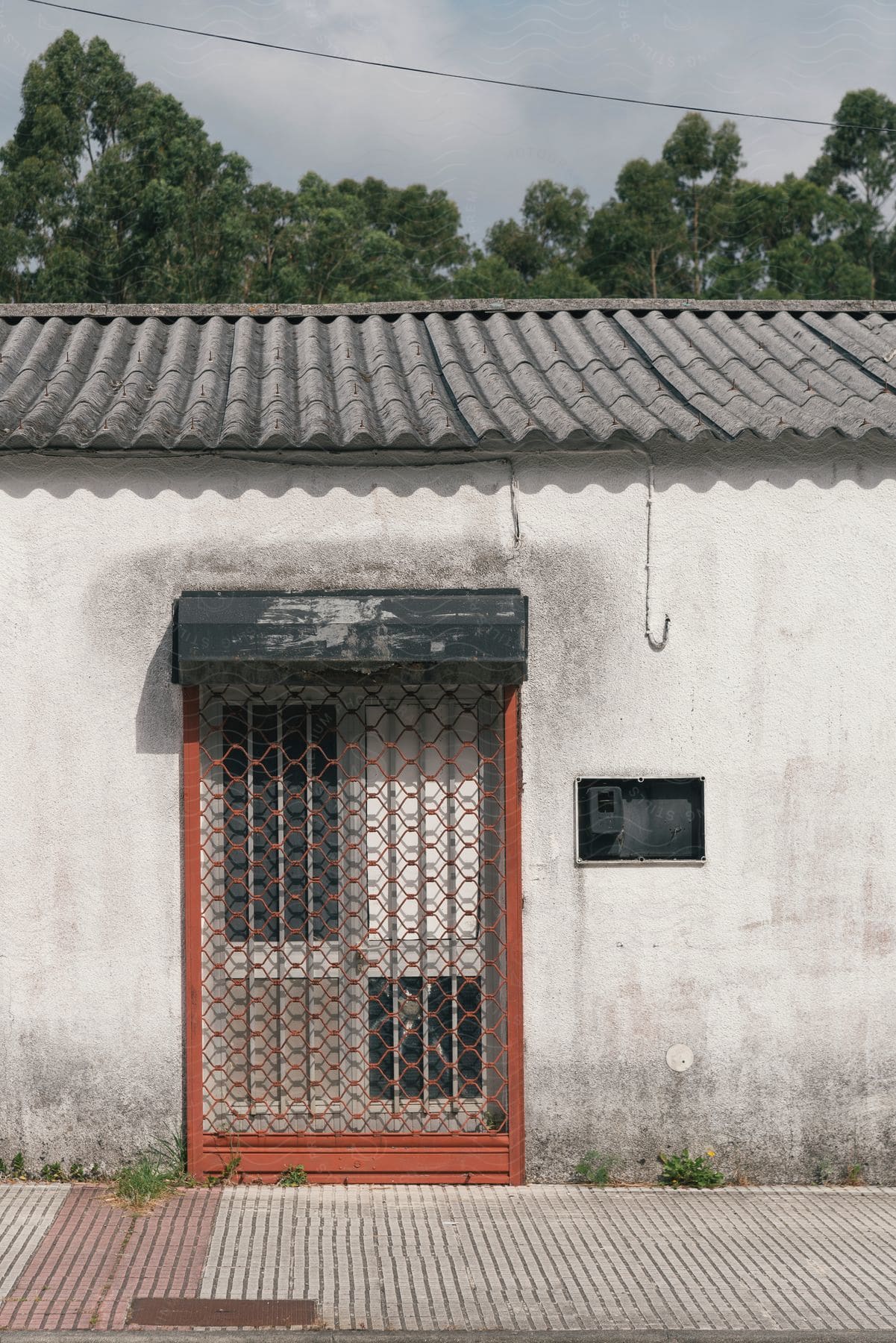 A worn-out house with a red door and a metal railing, under a cloudy sky. The building looks old, with a gray wall and traditional tiles on the roof.
