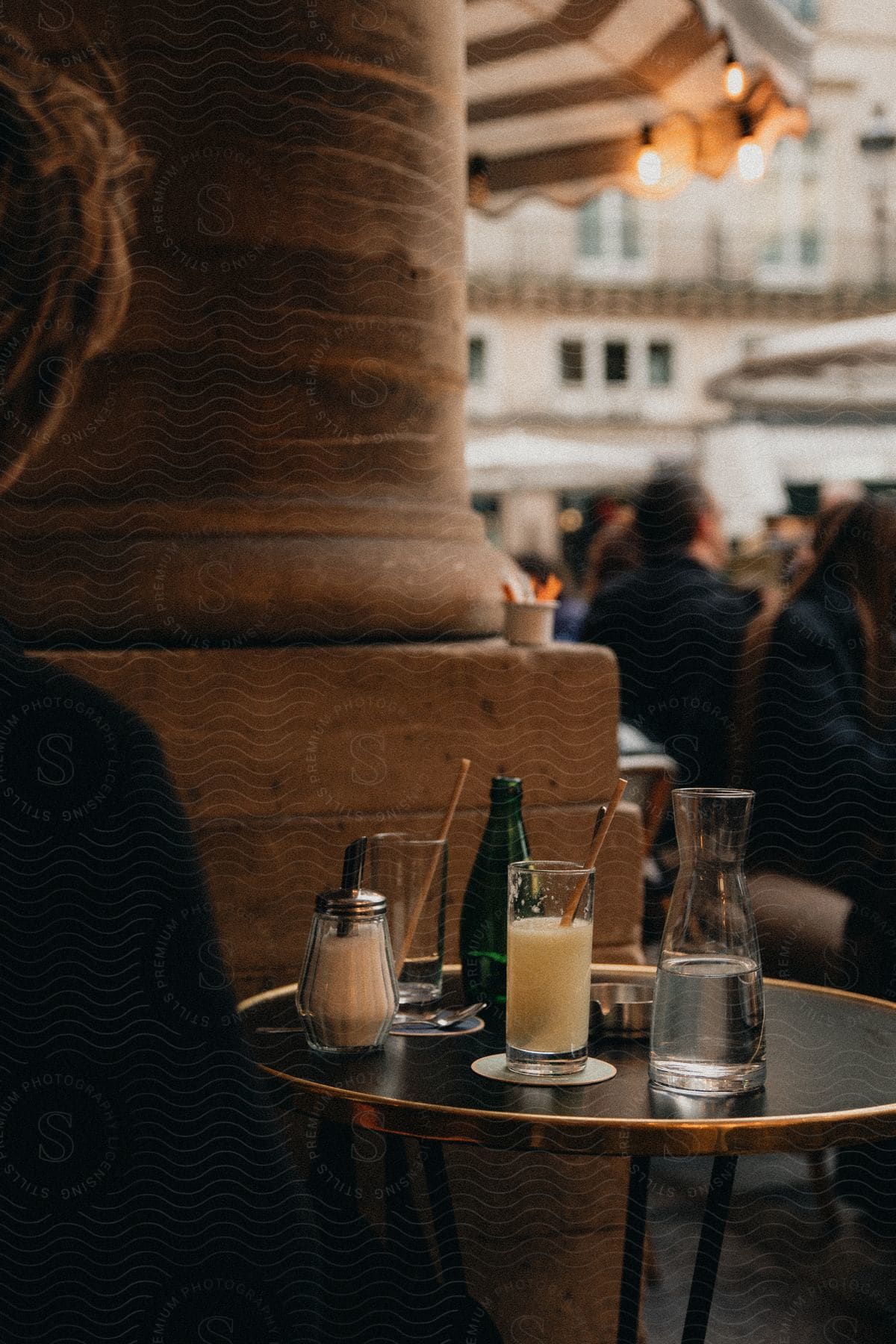Outdoor café table with a glass of juice, a bottle of water, and a sugar dispenser in front of a stone pillar, with blurred people in the background.
