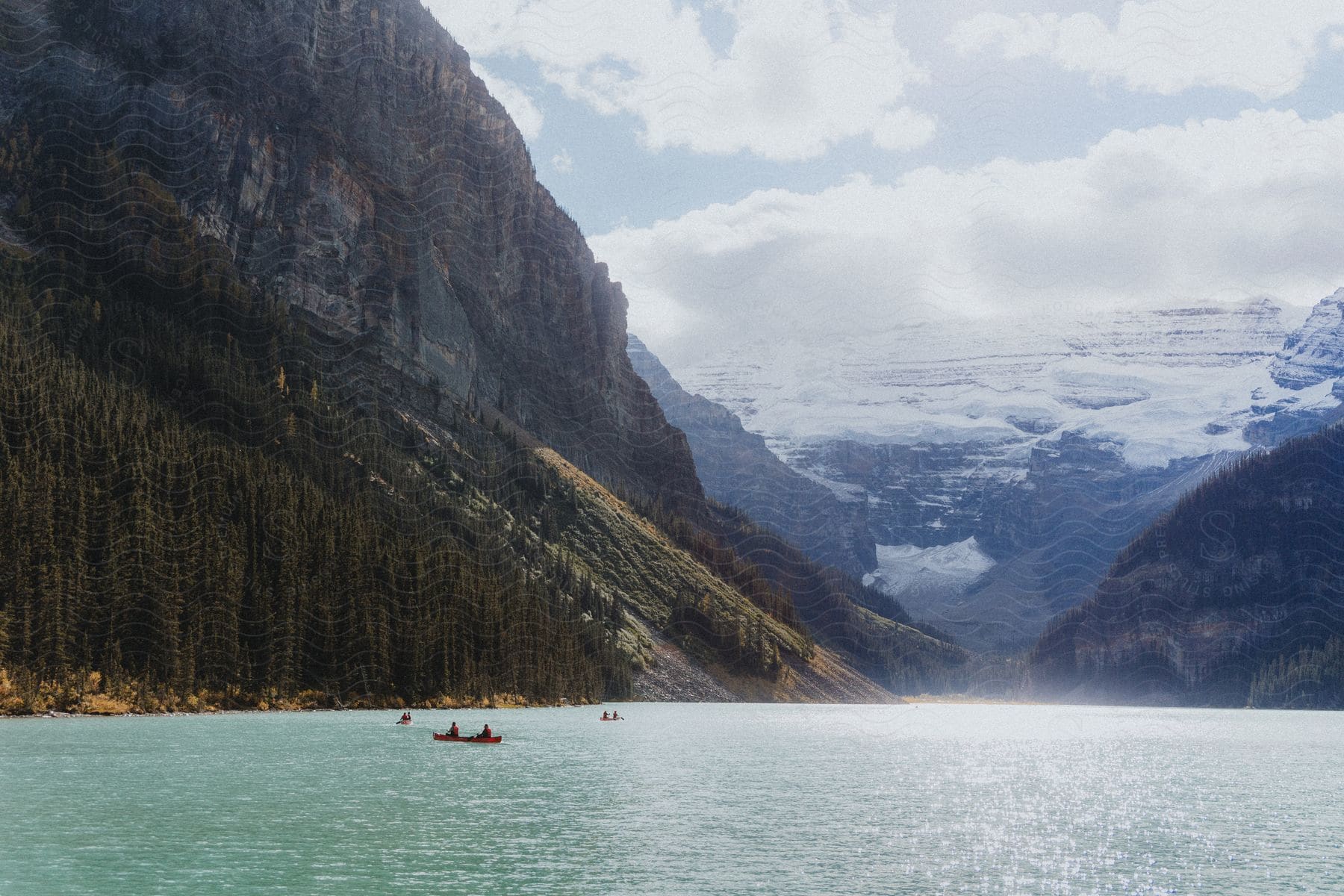 A tranquil scene of a turquoise lake surrounded by imposing mountains and dense forests. With canoes navigating the lake.