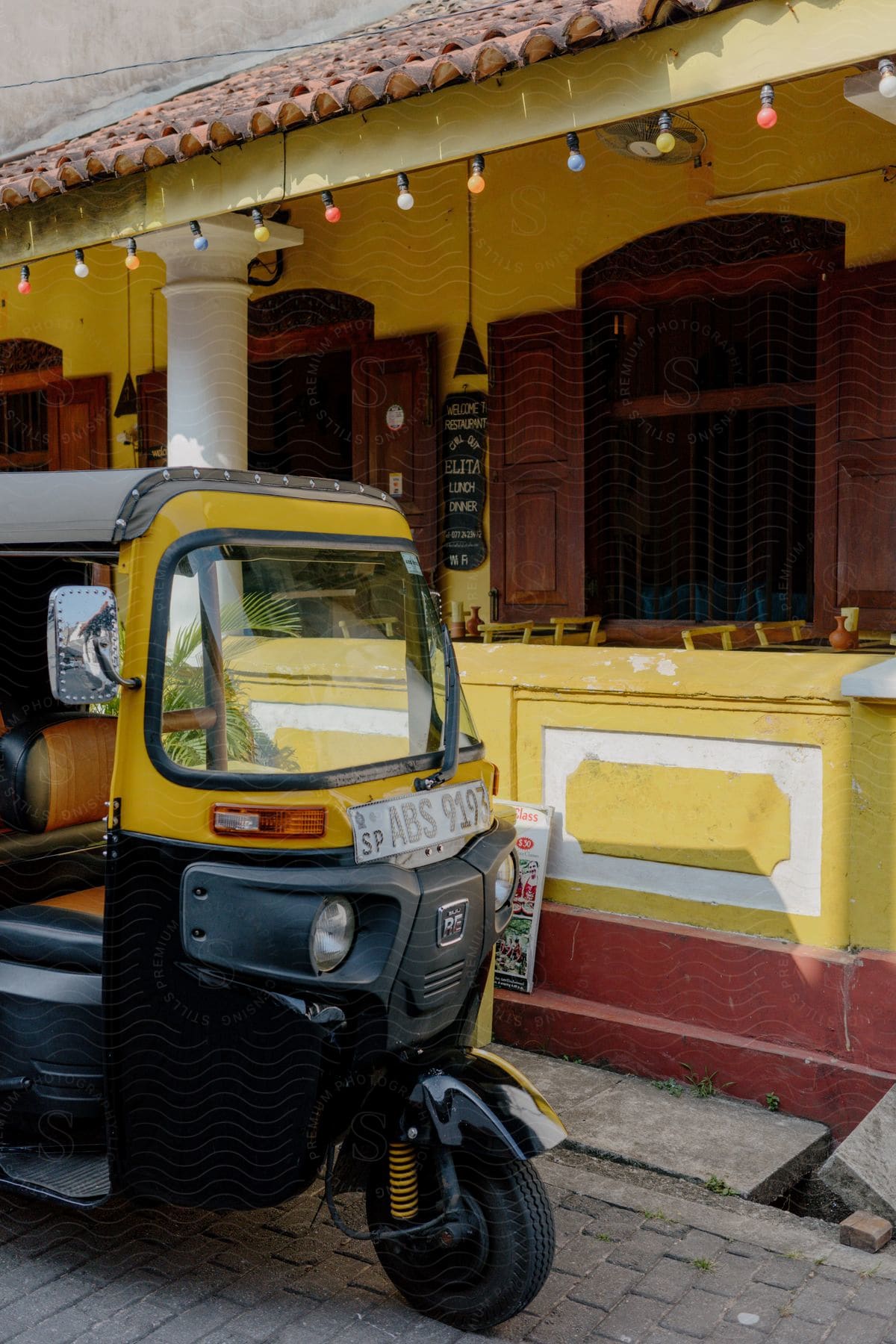 Three wheeled tuk tuk motor vehicle sits parked near restaurant balcony seating.