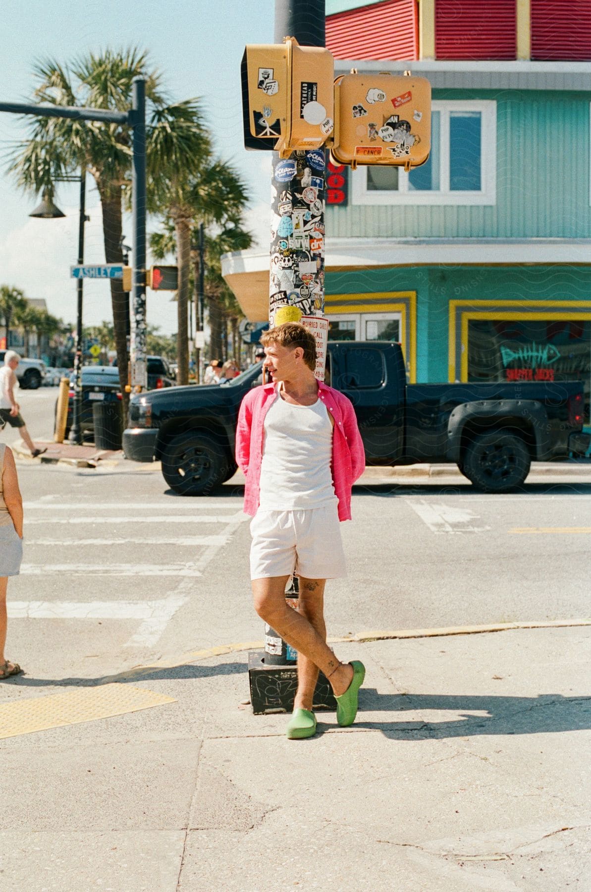 A man wearing white shorts and tee shirt with a pink shirt and green shoes leans against a traffic post