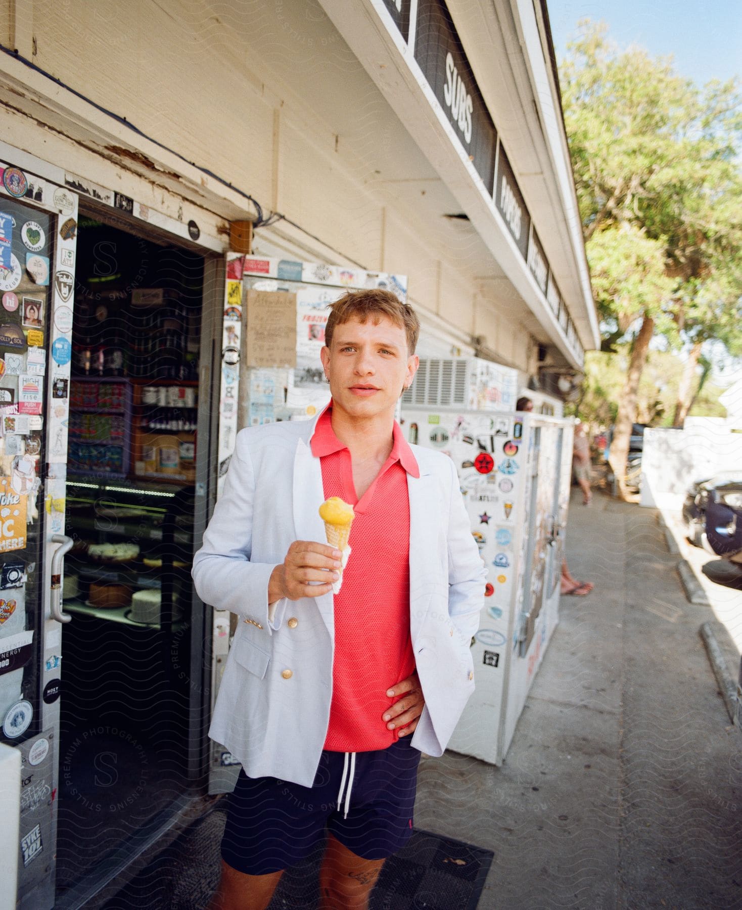Man with his hand on his waist wearing a white blazer and shorts while holding an ice cream next to a commercial establishment