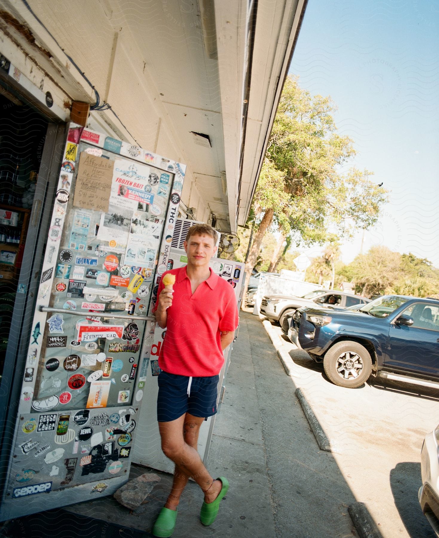 A man, clad in a red polo shirt, dark blue shorts, and green shoes, leans against a store's door while enjoying a cone of ice cream