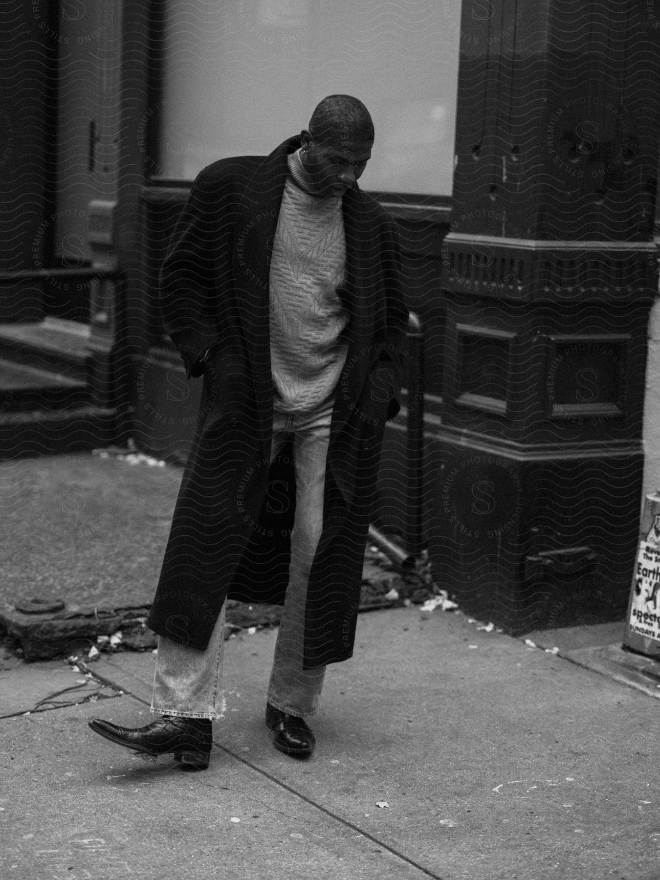 Person in long coat and jeans walking on a littered sidewalk by a dark building