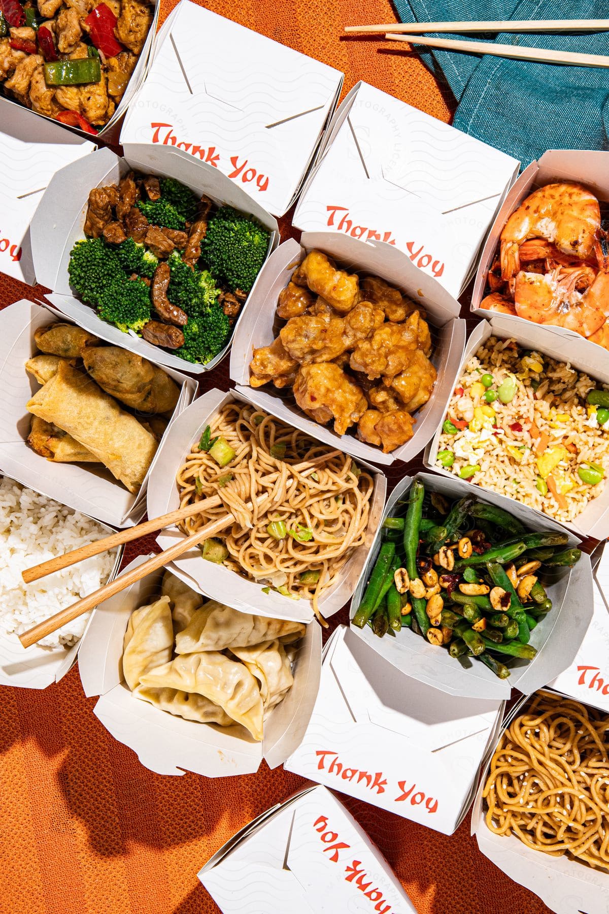 A table overflows with Chinese takeout boxes, chopsticks poised over a bowl of noodles and vegetables alongside a plate of meat.