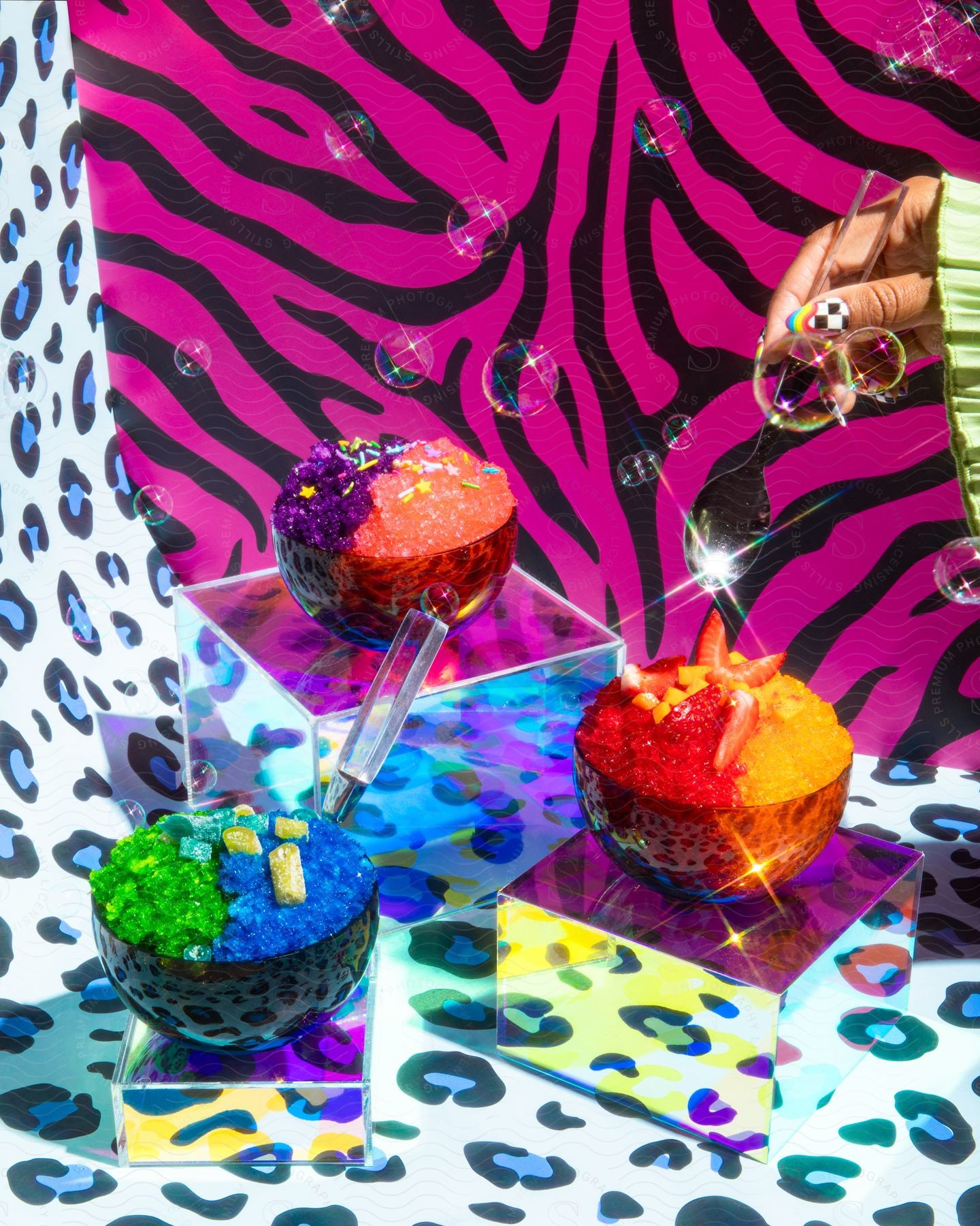 Three colorful shaved ice desserts in bowls on clear stands with a vibrant animal print background, a hand holding a spoon reaching towards one bowl.