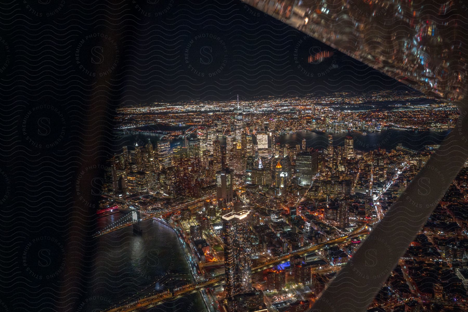 An aerial view from an airplane reveals New York City at night, with brightly lit skyscrapers lining the waterfront